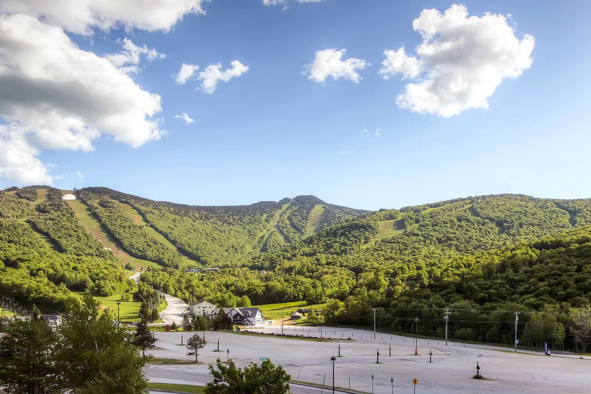 A sweeping view of the surrounding mountain range, green rolling hills, and a blue sky dappled with clouds. The image includes a road leading into the hills and an empty parking lot in the foreground, providing a sense of open space, nature, and adventure.