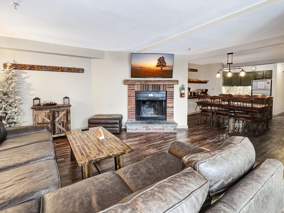 The image showcases a spacious, inviting living area featuring a crackling fireplace, a large sectional sofa perfect for group gatherings, a wooden coffee table ideal for snacks or games, and a TV displaying a scenic landscape. The dining area is visible in the background, suggesting space for shared meals and celebrations, all set in a warm, welcoming atmosphere.