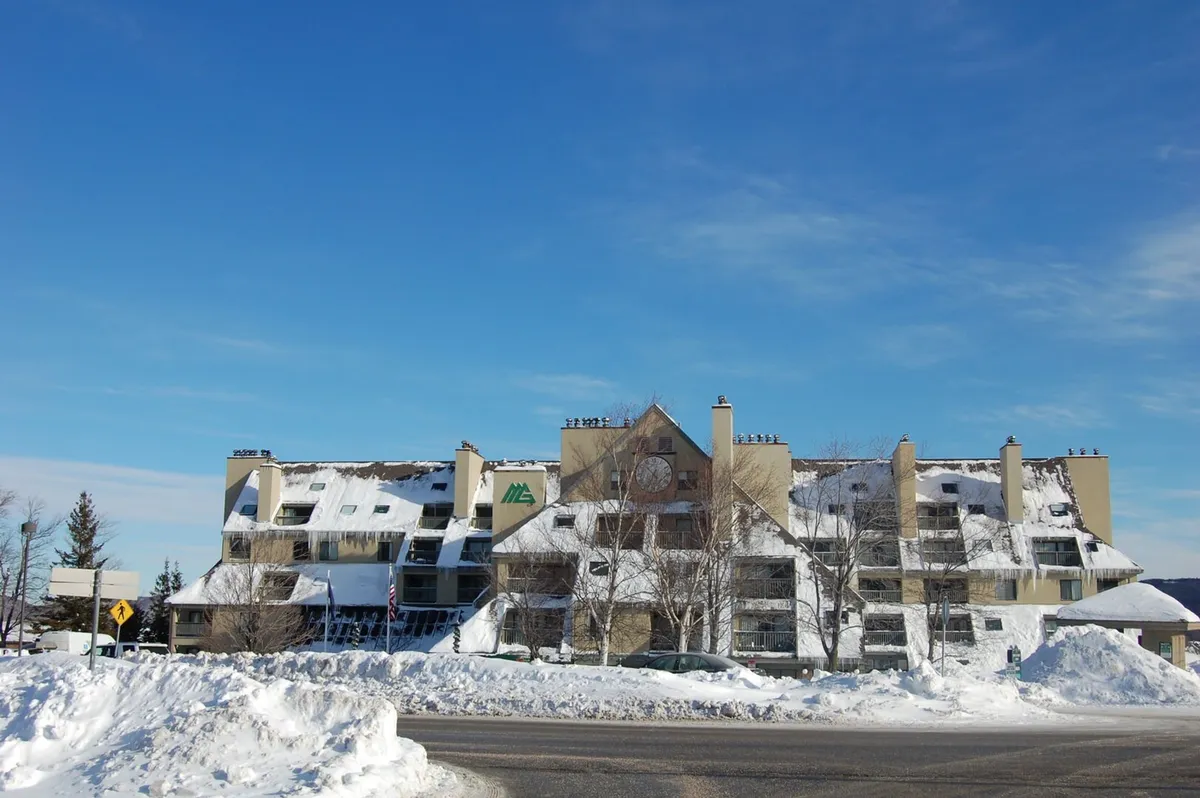 A snow-covered resort building under a crisp blue sky. Snow blankets the roof and grounds, while the facade invites guests in from a day on the slopes. The tranquil setting conveys both the excitement of winter activities and the comfort of returning to a cozy home base.
