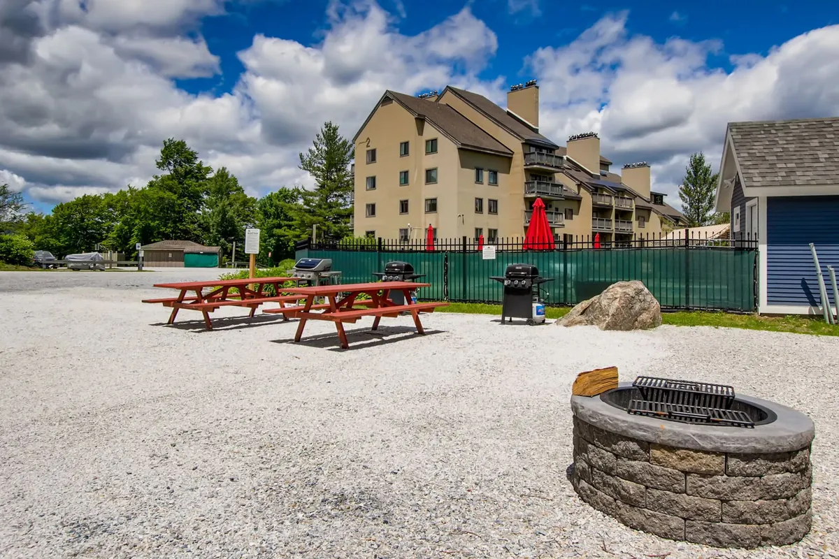 An exterior shot of the firepit and grill area, complete with picnic tables, a fire pit, grill stations, and the building in the background. The setting invites communal gatherings, barbecues, and sunset relaxation.
