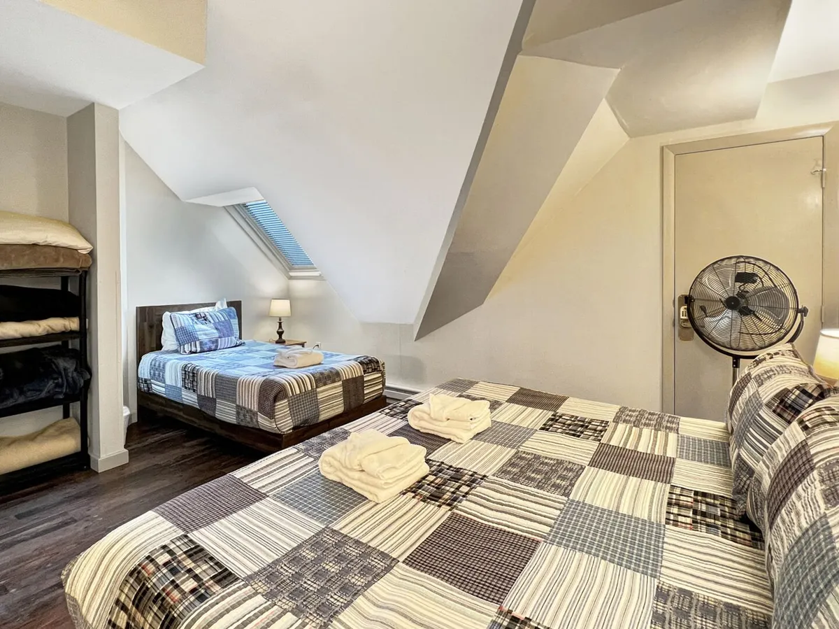 A family-friendly bedroom with a standard bed and an adjacent bunk bed, both dressed in soft linens. A lamp on the nightstand provides gentle lighting, and a fan sits in the corner for added comfort. The room feels welcoming for both adults and children.
