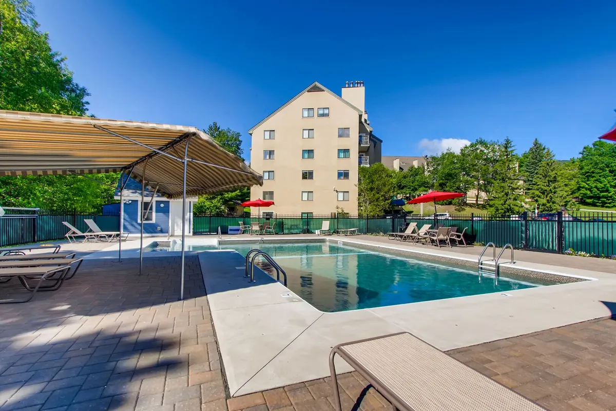 A spacious outdoor swimming pool area, surrounded by lounge chairs, umbrellas, and greenery, with the resort building in the background. The scene is set for both relaxation and social gatherings by the pool.