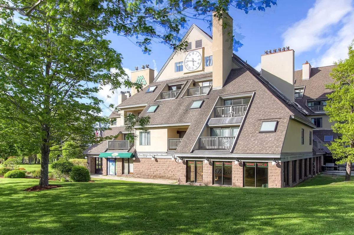 The main resort building with its distinctive clock tower, sloping roof, green lawn, and balconies. The image conveys a sense of arrival and the welcoming, communal nature of the property, set against a serene backdrop.