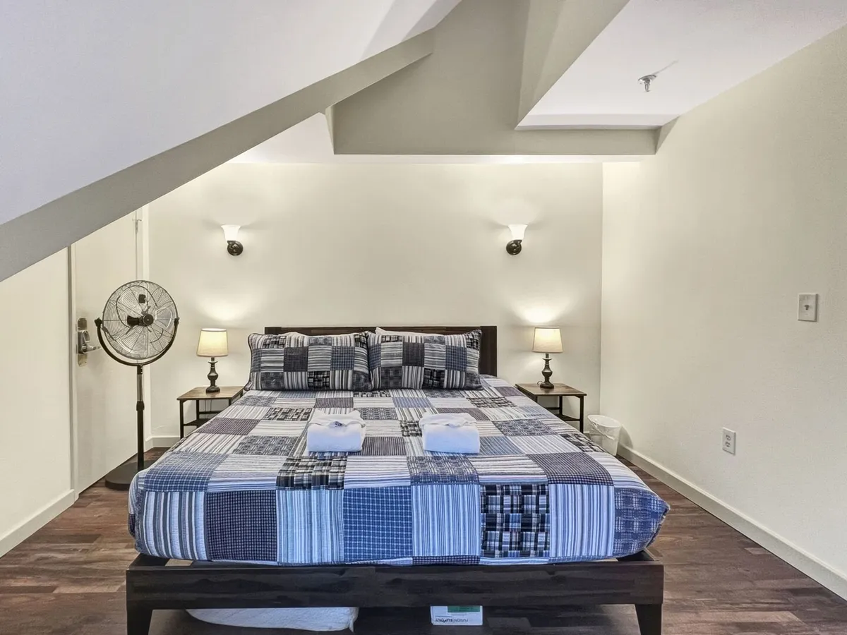 A cozy bedroom featuring a plush bed with patterned bedding, two bedside lamps casting warm light, wall-mounted fixtures, and a ceiling fan. The sunlight flows gently through the window, making the space feel inviting and restful.