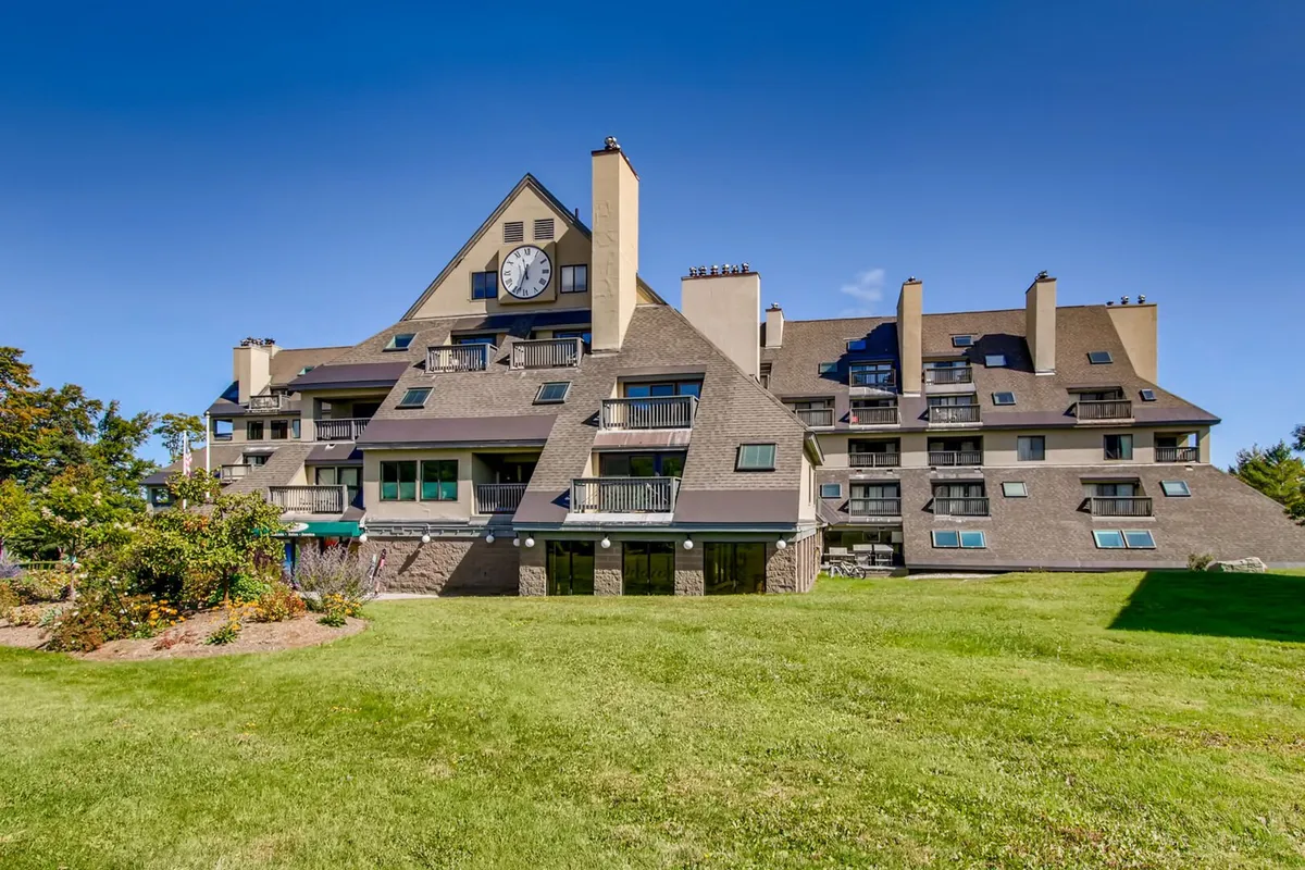 An exterior shot of the building with a large clock, green manicured lawn, and balconies with railings. The setting is serene, hinting at gatherings, sunset views, and enjoying the fresh mountain air outdoors or from the comfort of the balconies.