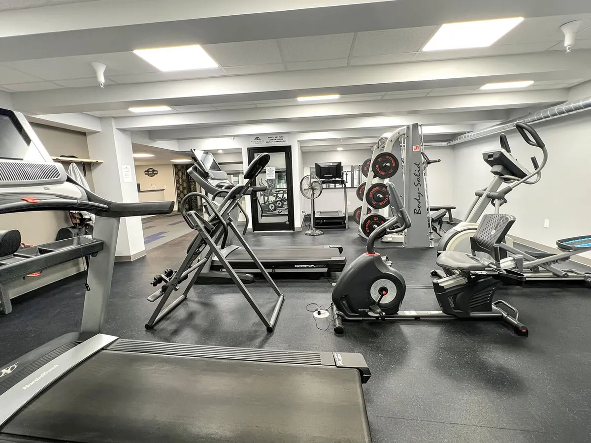 A well-equipped fitness center with treadmills, elliptical, weight machines, and a stationary bike is depicted. The space is light, organized, and ready for guests seeking exercise or personal challenges at any hour.