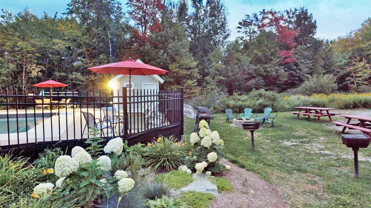 A vibrant outdoor pool scene with red umbrellas, a lush green lawn, colorful flower beds, and picnic tables. The pool is inviting and surrounded by spaces ideal for barbecuing, relaxing, and socializing in the sun.