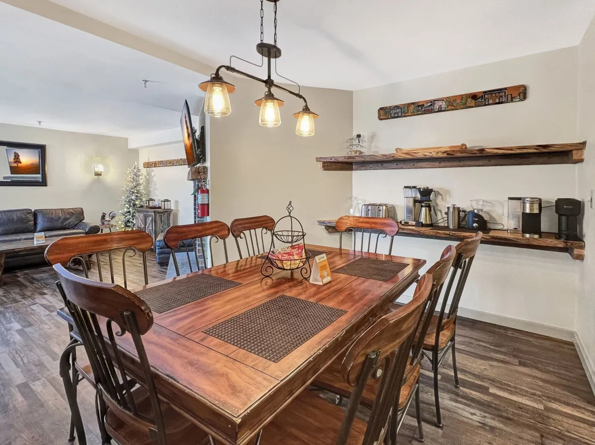 A stylish dining area with a spacious wooden table, modern hanging light fixtures, and shelving displaying kitchen items. A plush sofa is visible in the background, reinforcing the open flow between dining and living spaces. The space exudes warmth and is clearly designed for group meals and socializing.